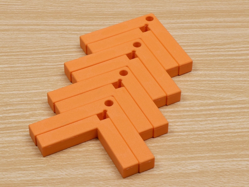 May include: Orange, L-shaped trivets arranged in a zig-zag pattern. Each trivet has a small hole in the corner. The trivets are made of a solid material and are resting on a light brown wooden surface.