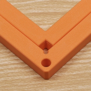 May include: An orange, right-angle corner tool, likely for woodworking or construction. The tool is made of a solid, textured material and features a hole for hanging or securing. The tool is positioned on a light-colored wooden surface.