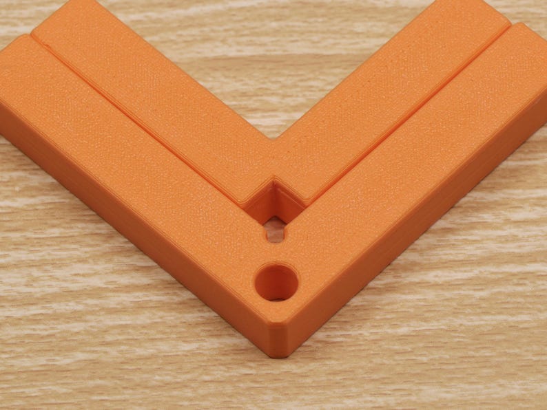 May include: An orange, right-angle corner tool, likely for woodworking or construction. The tool is made of a solid, textured material and features a hole for hanging or securing. The tool is positioned on a light-colored wooden surface.