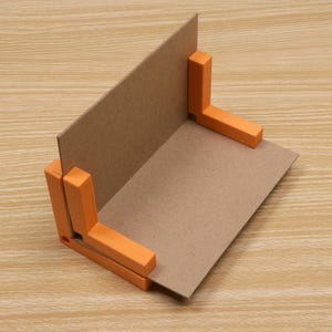May include: A cardboard corner clamp with orange plastic supports. The clamp is designed to hold two pieces of cardboard at a 90-degree angle. The background is a light brown wooden surface.