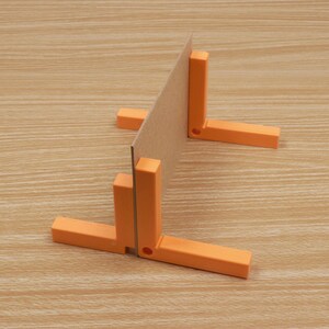 May include: An orange right angle tool with a cardboard insert, set against a light wood grain surface. The tool is composed of two perpendicular arms and a vertical support, designed to hold the cardboard at a 90-degree angle. The tool is likely used for woodworking or crafts.