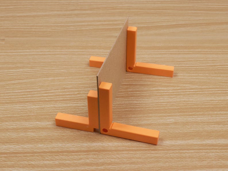 May include: An orange right angle tool with a cardboard insert, set against a light wood grain surface. The tool is composed of two perpendicular arms and a vertical support, designed to hold the cardboard at a 90-degree angle. The tool is likely used for woodworking or crafts.