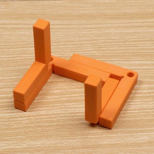 May include: An orange, foldable display stand. The stand is made of rectangular blocks and has a simple, geometric design. It is designed to hold items for display. The stand is on a light brown wooden surface.