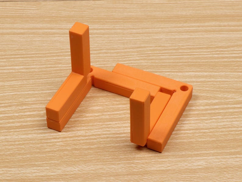 May include: An orange, foldable display stand. The stand is made of rectangular blocks and has a simple, geometric design. It is designed to hold items for display. The stand is on a light brown wooden surface.