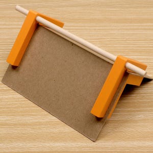 May include: A small, handmade wooden jig with orange plastic clamps holding a piece of brown cardboard. A light-colored wooden dowel is held in place by the clamps. The jig is designed for crafting or small projects.