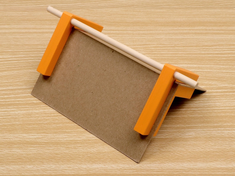 May include: A small, handmade wooden jig with orange plastic clamps holding a piece of brown cardboard. A light-colored wooden dowel is held in place by the clamps. The jig is designed for crafting or small projects.