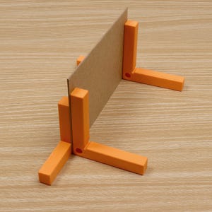 May include: A cardboard sheet is held upright by an orange plastic right-angle bracket. The bracket has three legs and a slot to hold the cardboard. The background is a light brown wood grain.