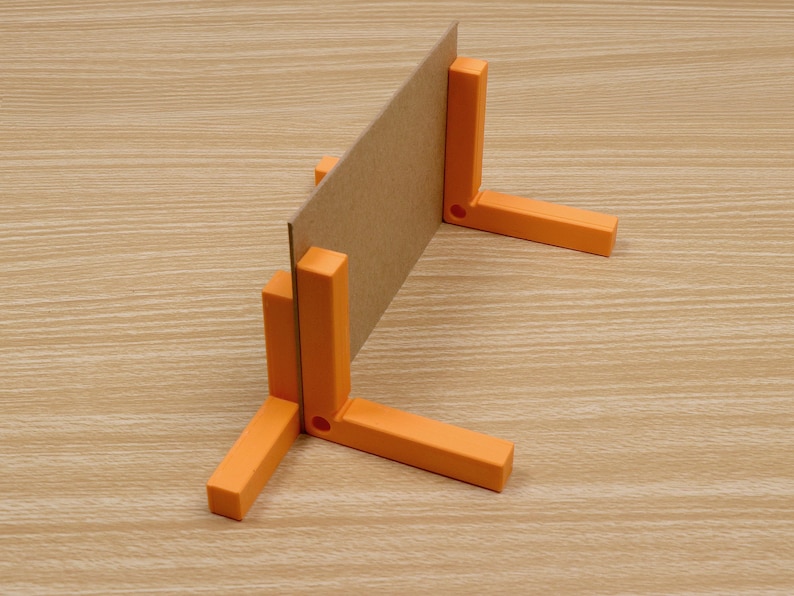 May include: A cardboard sheet is held upright by an orange plastic right-angle bracket. The bracket has three legs and a slot to hold the cardboard. The background is a light brown wood grain.