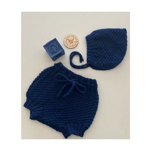 May include: A blue knitted baby bonnet and matching bloomers. The bonnet has a long tie that can be tied under the chin. The bloomers have a drawstring waist.