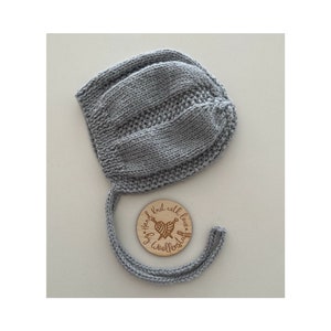 May include: Grey knitted baby bonnet with a tie, hand-knitted with love by Woollenstuff. The tag is a round wooden circle with the text "Hand Knit with love by Woollenstuff" and a heart with knitting needles.