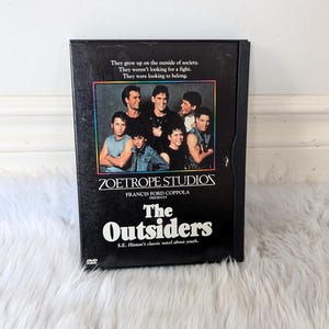 May include: A black DVD case for the movie "The Outsiders" by Zoetrope Studios. The case features a photo of the cast and the film's title in white. The cover also includes the text "They grew up on the outside of society."