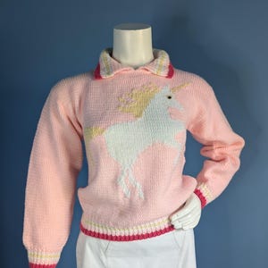 May include: A long-sleeved, light pink knit sweater featuring a white unicorn graphic with a yellow mane and horn. The sweater has a collared neckline and ribbed cuffs and hem with horizontal stripes in pink, yellow, and white colors.