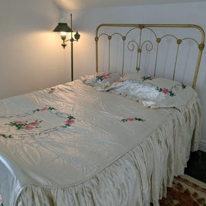 May include: A vintage bed with a gold-colored metal frame and a cream-colored satin bedspread. The bedspread and pillowcases are embroidered with pink roses and green leaves. A matching ruffled bed skirt completes the set. A floor lamp with a green shade is in the background.