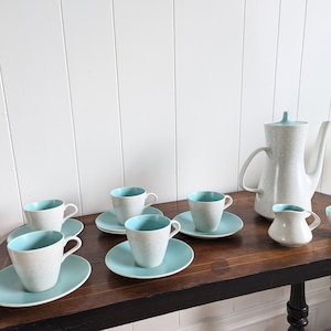 Puede incluir: Un juego de tazas de té vintage y una tetera con un esmalte blanco y azul moteado. Las tazas y la tetera tienen un borde azul claro y están dispuestas sobre una mesa de madera.