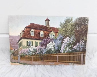May include: A vintage postcard featuring a photo of a house with a red roof and a fence in front of it. The house is surrounded by a lush garden with purple and white flowers.