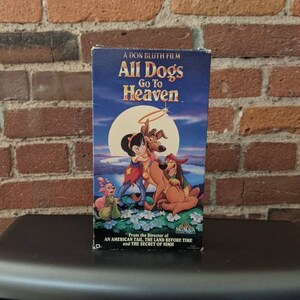 May include: A VHS tape for the animated film "All Dogs Go To Heaven". The cover features a blue sky with a full moon and a halo above a dog and a girl. The text "A Don Bluth Film" is at the top of the cover, and the text "From the Director of An American Tail, The Land Before Time and The Secret of Nimh" is at the bottom.