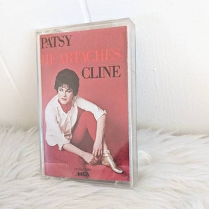 May include: A vintage cassette tape with a red background featuring Patsy Cline. The text "Patsy Cline" and "Heart Aches" are printed in black on the front of the cassette tape. The cassette tape is in a clear plastic case.