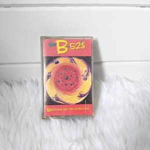 May include: A cassette tape with a colourful illustration of a swirling vortex with purple, yellow and red colours. The text "the B-52's" is in yellow and red on the cassette tape. The text "Bouncing Off the Satellites" is in yellow on the cassette tape.