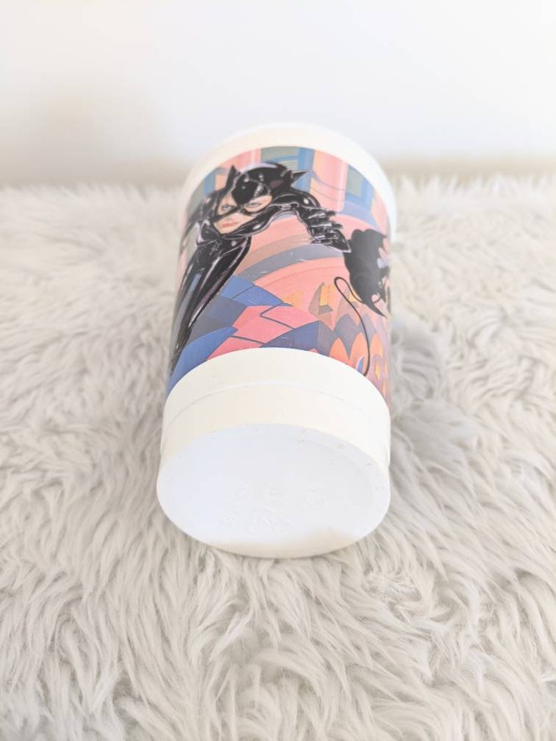 May include: White plastic cup with a colorful image of Catwoman from the Batman movie. The image features Catwoman in a black leather suit with a whip and a cityscape background.