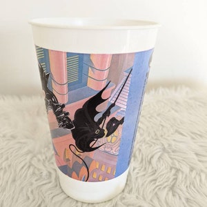May include: White plastic cup with a colorful graphic of a black figure climbing a building. The graphic is a stylized cityscape with pink, blue, and yellow colors.