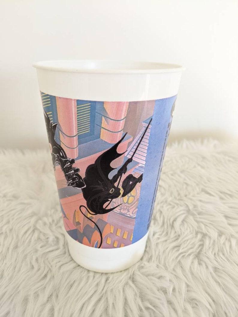 May include: White plastic cup with a colorful graphic of a black figure climbing a building. The graphic is a stylized cityscape with pink, blue, and yellow colors.