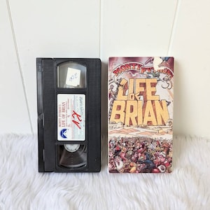 Może przedstawiać: Kaseta VHS z filmem Monty Python's Life of Brian. Kaseta jest czarna z przezroczystym okienkiem z plastiku, które pokazuje tytuł filmu i scenę z filmu. Kaseta leży obok okładki filmu, na której znajduje się rysunkowa ilustracja tłumu ludzi.