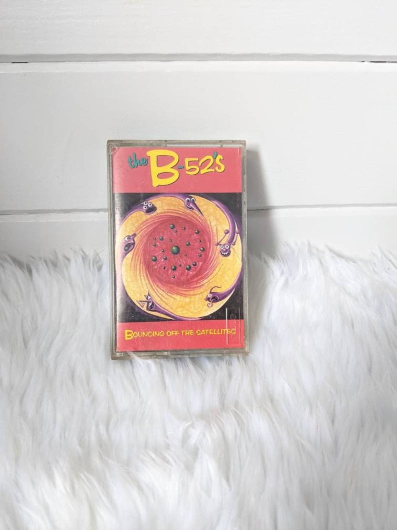 May include: A cassette tape with a colourful illustration of a swirling vortex with purple, yellow and red colours. The text "the B-52's" is in yellow and red on the cassette tape. The text "Bouncing Off the Satellites" is in yellow on the cassette tape.