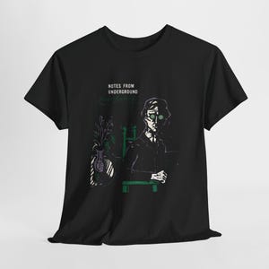 以下が含まれることがあります： 黒いTシャツ。メガネをかけた人物、花瓶、そして「NOTES FROM UNDERGROUND」と「Fyodor Dostoevsky」の文字が描かれたグラフィックイラスト。デザインは緑、白、黒を使用しています。