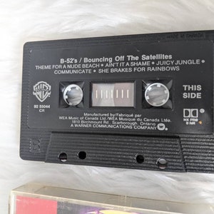May include: Black cassette tape with the title "B-52's / Bouncing Off The Satellites" and the song titles "Theme For A Nude Beach", "Ain't It A Shame", "Juicy Jungle", "Communicate", and "She Brakes For Rainbows". The cassette tape is labelled "This Side" and has a Warner Bros. Records logo.