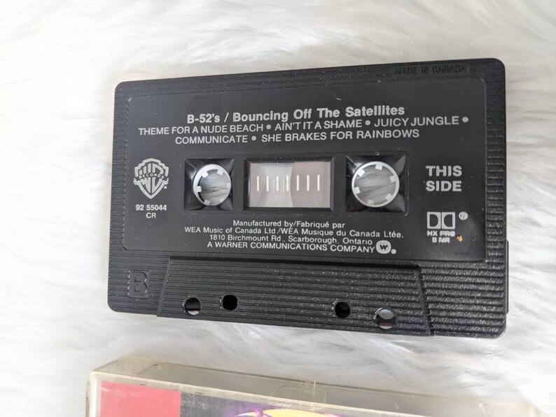 May include: Black cassette tape with the title "B-52's / Bouncing Off The Satellites" and the song titles "Theme For A Nude Beach", "Ain't It A Shame", "Juicy Jungle", "Communicate", and "She Brakes For Rainbows". The cassette tape is labelled "This Side" and has a Warner Bros. Records logo.