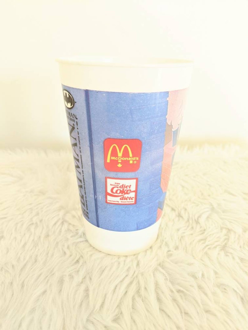 May include: White plastic cup with a blue and pink Batman Returns design. The cup features the McDonald's logo and a Diet Coke logo.