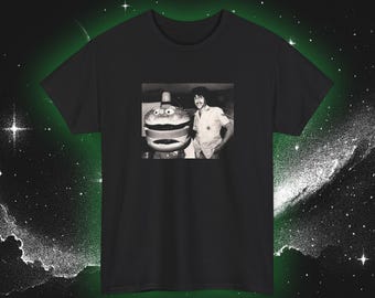May include: Black t-shirt featuring a black and white image of a man in a uniform standing next to a large hamburger statue. The image is centered on the shirt, with a dark green and white space-themed background.