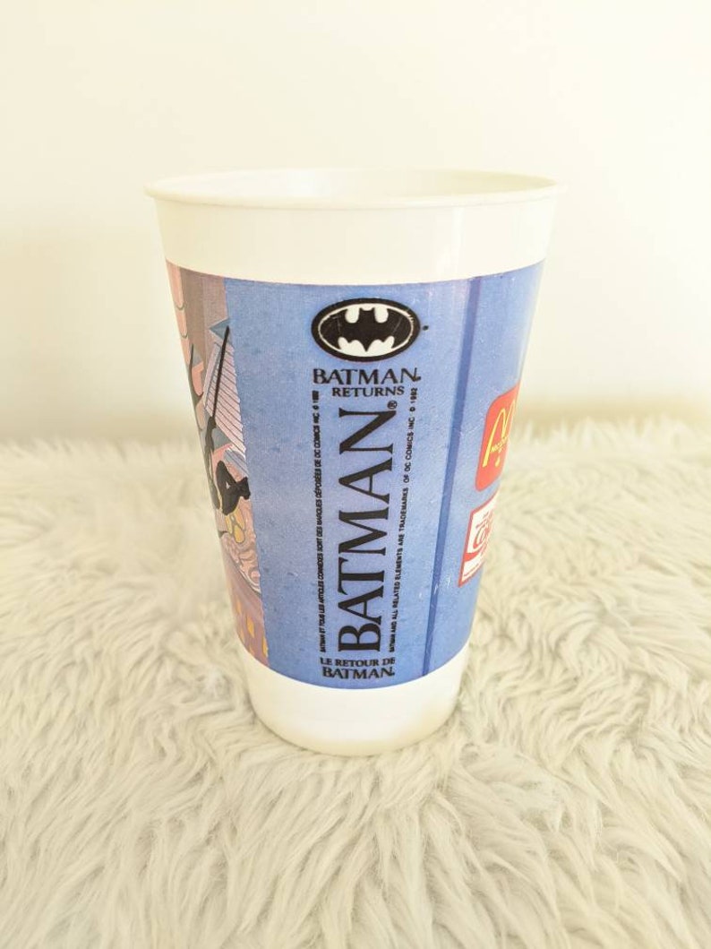 May include: A white plastic cup with a blue and red Batman Returns design. The cup features the Batman logo and the text "Batman Returns" and "Le Retour de Batman".