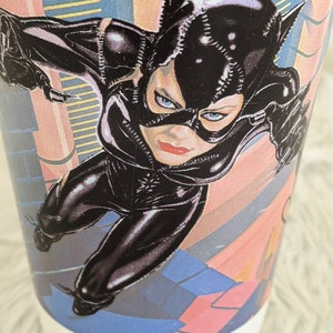 May include: A black and white illustration of Catwoman, a fictional superhero, wearing a black leather suit and mask. She is posed in a dynamic action pose with her arms outstretched. The background is a colorful abstract design.