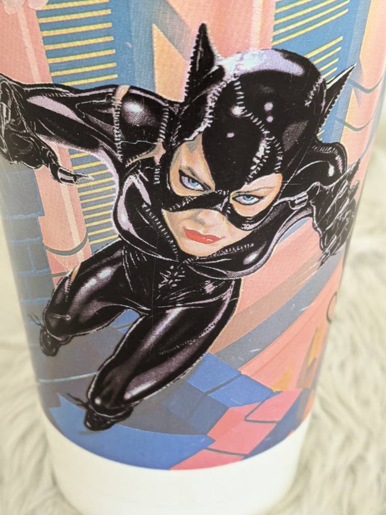 May include: A black and white illustration of Catwoman, a fictional superhero, wearing a black leather suit and mask. She is posed in a dynamic action pose with her arms outstretched. The background is a colorful abstract design.