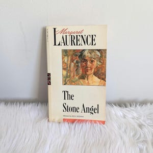 Peut inclure: Un livre de poche intitulé "The Stone Angel" de Margaret Laurence. La couverture présente un tableau d'un visage de femme.