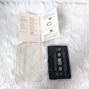May include: Black cassette tape with a white label. The label has the text "Theme Forcen - Off The Satellites - Juicy Jungle" and "This Side".