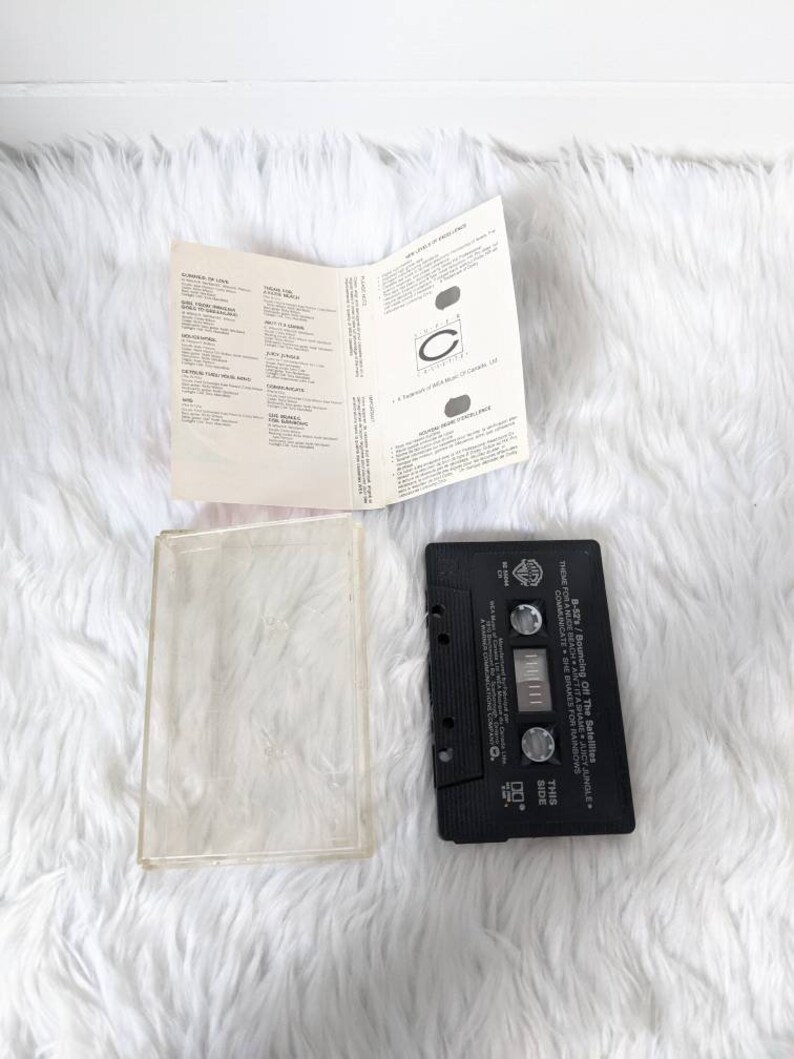 May include: Black cassette tape with a white label. The label has the text "Theme Forcen - Off The Satellites - Juicy Jungle" and "This Side".