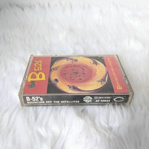 May include: A cassette tape with a colourful, swirling design on the front. The tape is titled "B-52's Bouncing Off The Satellites" and has the Warner Bros. Records logo.
