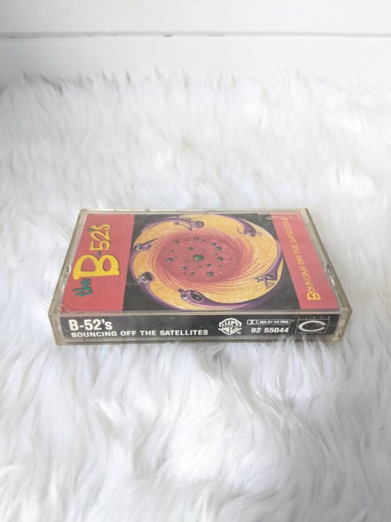 May include: A cassette tape with a colourful, swirling design on the front. The tape is titled "B-52's Bouncing Off The Satellites" and has the Warner Bros. Records logo.