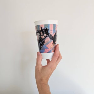 May include: White plastic cup with a colorful image of Catwoman in a black leather suit, leaping through a building with a whip. The background is a pink, blue, and green cityscape.