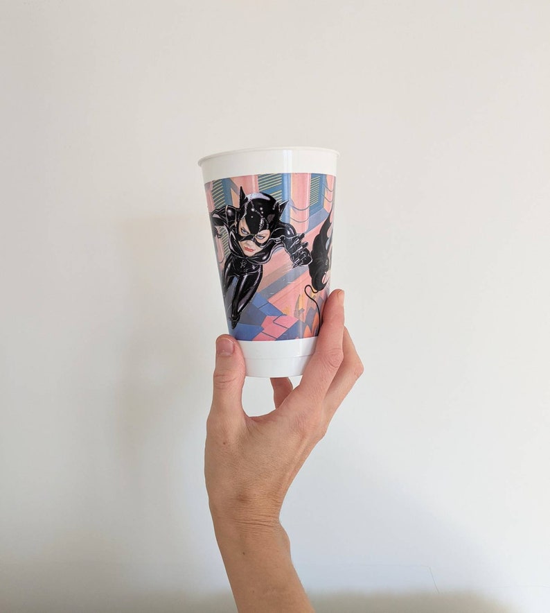 May include: White plastic cup with a colorful image of Catwoman in a black leather suit, leaping through a building with a whip. The background is a pink, blue, and green cityscape.