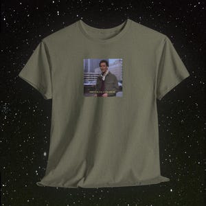 May include: Olive green t-shirt featuring a small image of a man with the text "PRETTY BLEAK, IF YOU ASK ME." The image is centered on the front of the shirt. The shirt has short sleeves and a crew neck.
