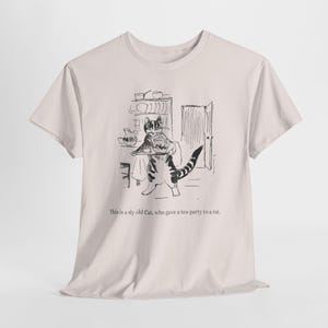 Puede incluir: Camiseta beige claro con una ilustración en blanco y negro de un gato sosteniendo una bandeja. El gato está en un entorno de cocina. El texto "This is a sly old Cat, who gave a tea-party to a rat." está debajo.