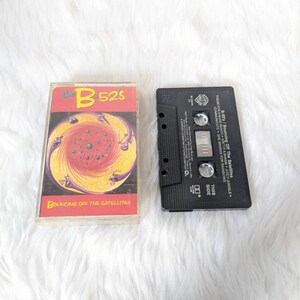 May include: A cassette tape of the B-52's album "Bouncing Off The Satellites". The cassette tape is black with a white label. The album cover features a colourful, psychedelic design with a swirling pattern and a black and white image of a bird.
