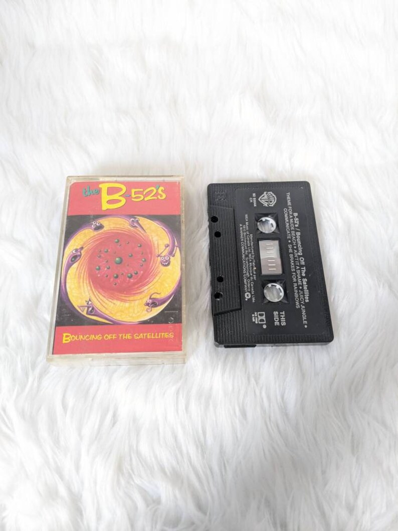 May include: A cassette tape of the B-52's album "Bouncing Off The Satellites". The cassette tape is black with a white label. The album cover features a colourful, psychedelic design with a swirling pattern and a black and white image of a bird.