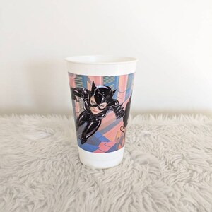 May include: A white plastic cup with a colorful image of Catwoman from the Batman movie. The image shows Catwoman in a black leather suit with a whip.