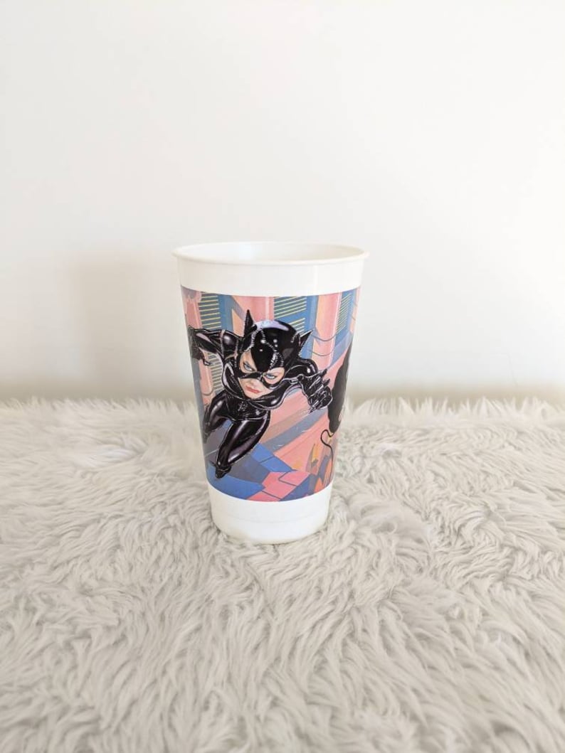 May include: A white plastic cup with a colorful image of Catwoman from the Batman movie. The image shows Catwoman in a black leather suit with a whip.