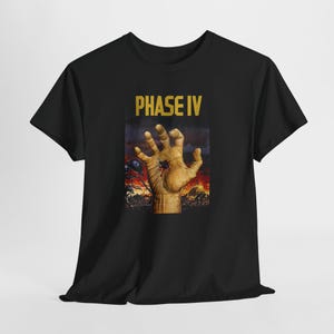 Op de afbeelding: Zwart T-shirt met een afbeelding uit de film "Phase IV". Het ontwerp toont een hand met een gat in de palm, tegen een vurige, apocalyptische achtergrond. De woorden "PHASE IV" staan in gouden letters boven de hand.