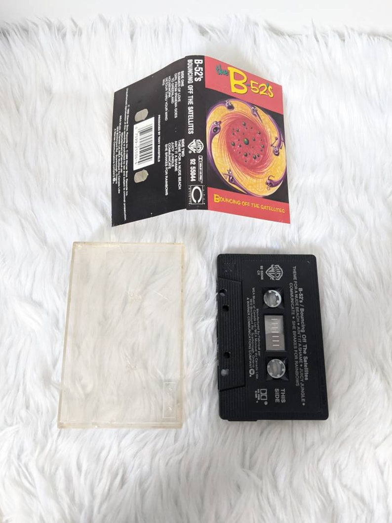 May include: A black cassette tape case with the title "B-52's Bouncing Off The Satellites" printed on the front. The case has a colourful image of a swirling vortex with black, red, yellow, and orange colours.